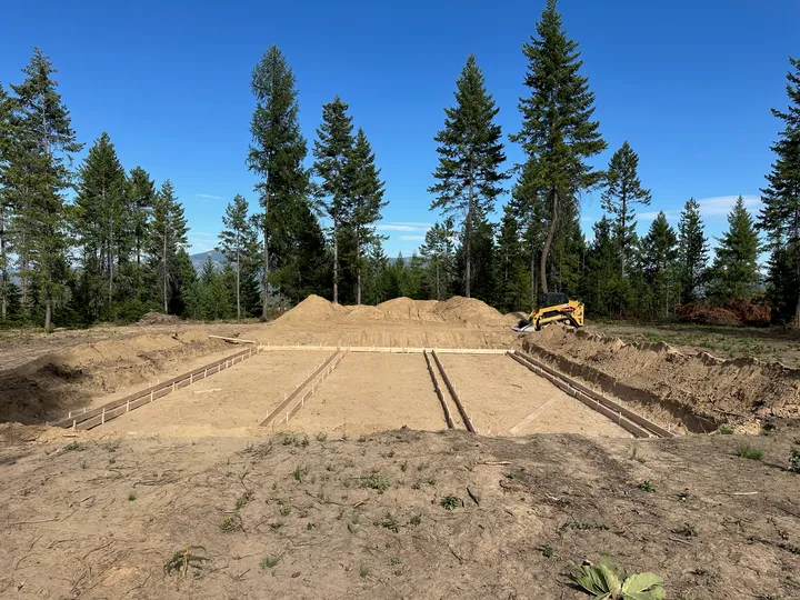 Staked and graded foundation pad on a cleared lot with ponderosa pines and blue sky near Newport, WA